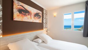 Una habitación de hotel con una cama grande, un cuadro de un ojo en la pared, vista al mar y un papel tapiz estampado.