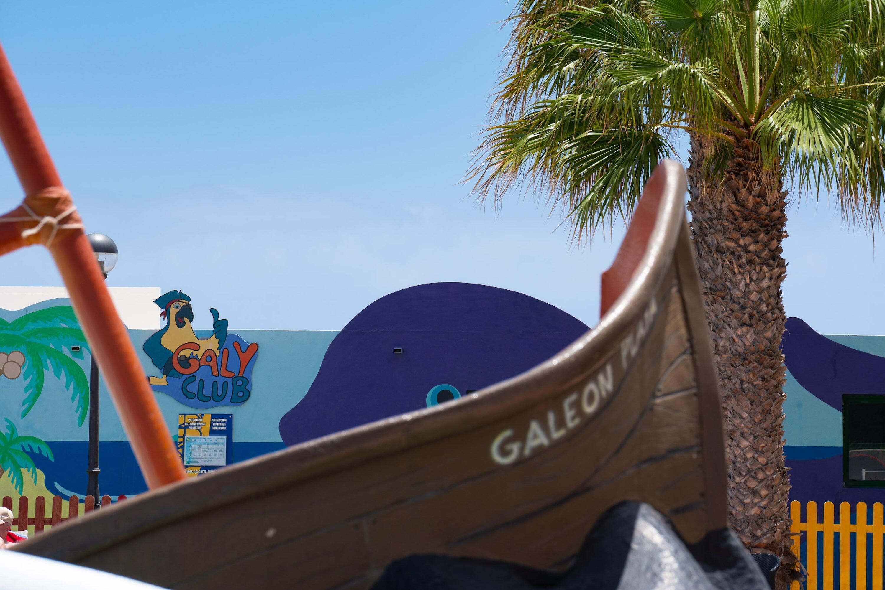 Een boot met de tekst 'GALEON PALACE' op de zijkant, vlakbij een palmboom en een gebouw met een muurschildering van een haan en de tekst 'GALY CLUB'.