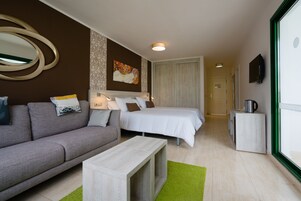 Habitación de hotel con un sofá gris, una cama con ropa de cama blanca, una pequeña mesa de madera y una alfombra verde.