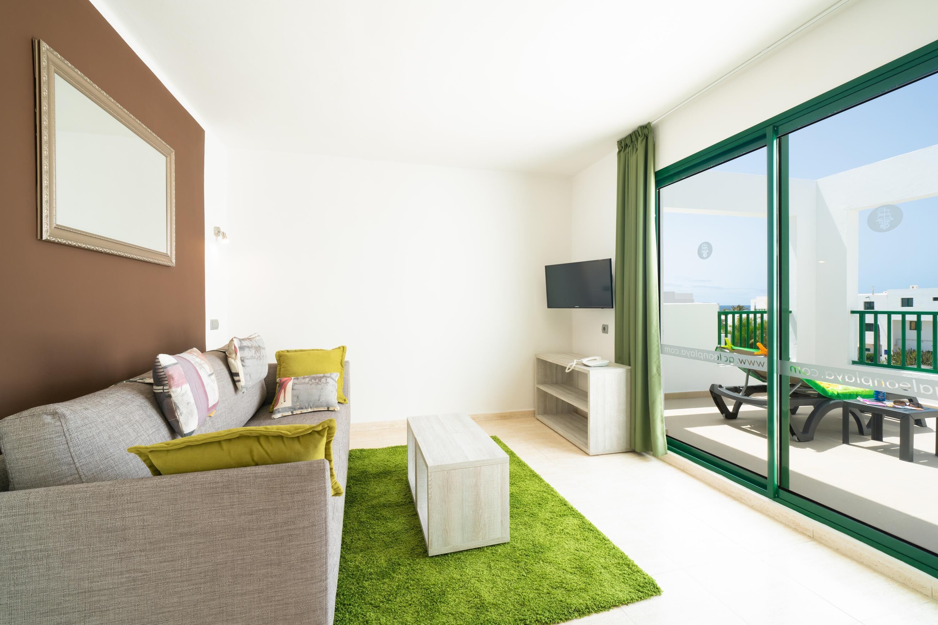 A modern living room with a grey sofa, a green rug, a flat-screen TV, and a balcony with outdoor furniture.