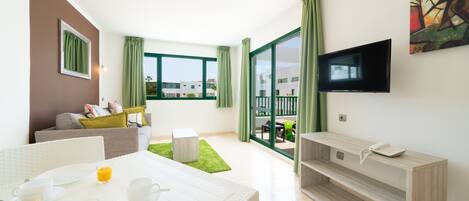 A modern hotel room with a dining area, a sofa, a TV mounted on the wall, and a balcony with a view of buildings.