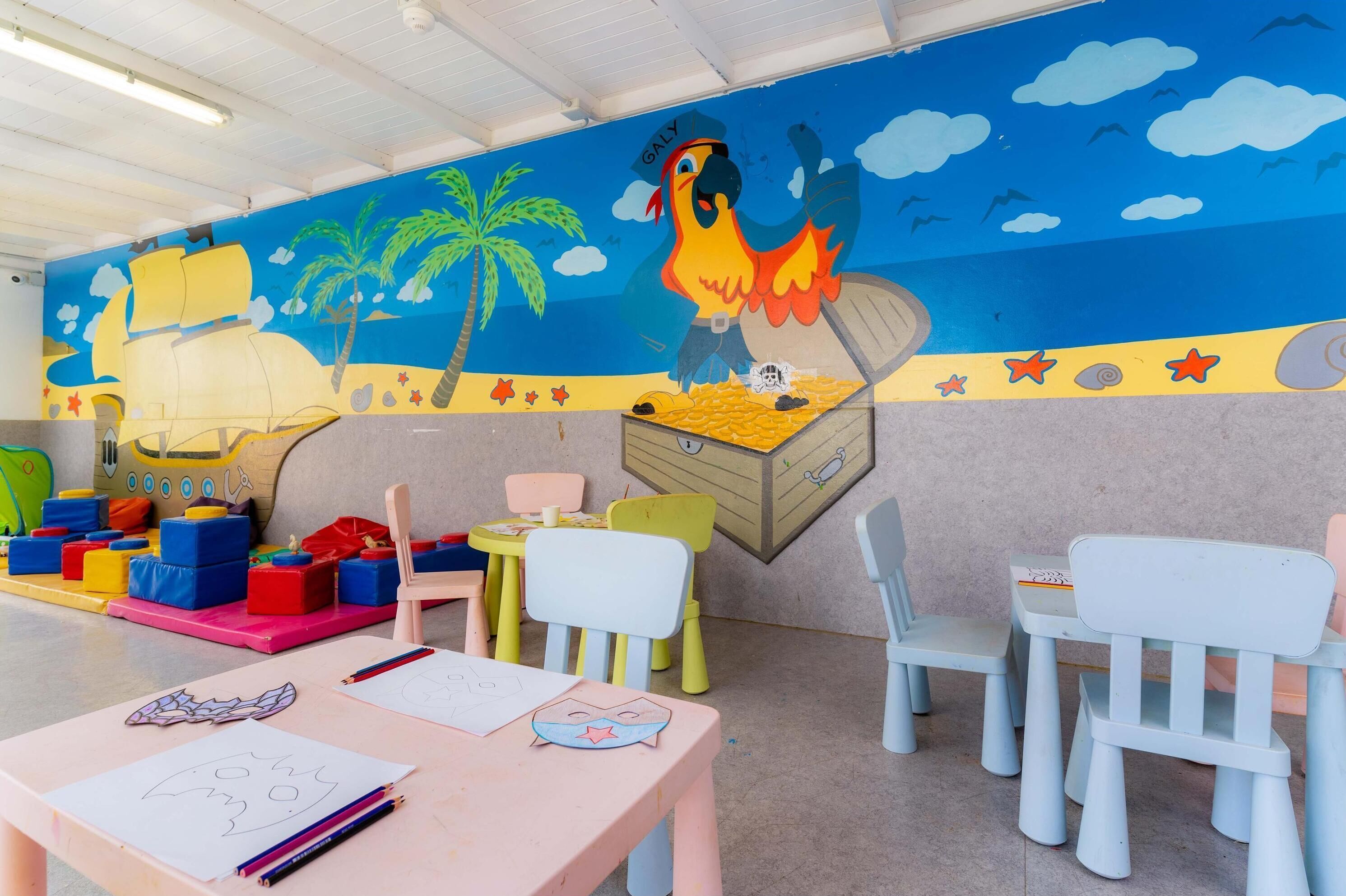 Zona de juegos infantiles con mobiliario de colores y un mural de un loro y un barco pirata.