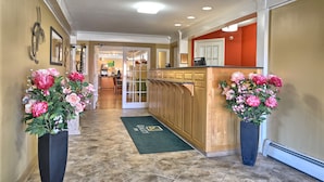 Un vestíbulo de hotel con mostrador de recepción, dos grandes arreglos florales, una alfombra de entrada verde y vista al comedor.