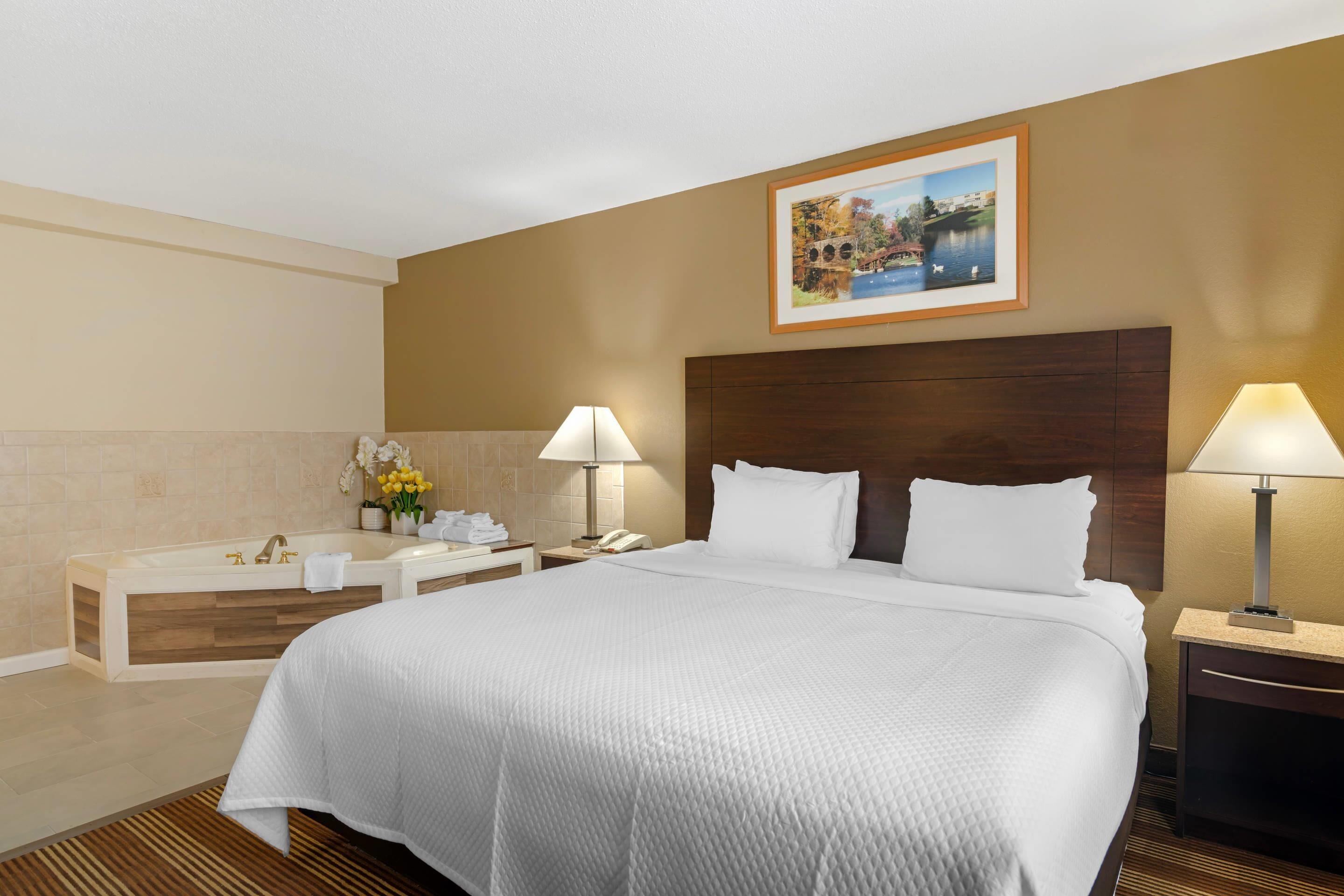 Ein Hotelzimmer mit einem großen Bett, einem hölzernen Kopfteil, zwei Nachttischen mit Lampen, einem Beistelltischchen mit Telefon sowie einem Badezimmer mit Badewanne und einem gerahmten Bild an der Wand.