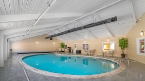 Una piscina interior con suelo de baldosas, techo con conductos visibles y una zona de estar con sillas y una mesa.