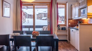 A dining area with a table set for two, a window with curtains, and a view of buildings and snow outside. - Pierre & Vacances Residence Le Quartz (La Plagne-Tarentaise)