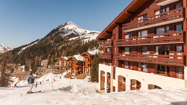A ski resort with wooden buildings, a mountain in the background, and a person skiing. - Pierre & Vacances Residence Le Quartz (La Plagne-Tarentaise)