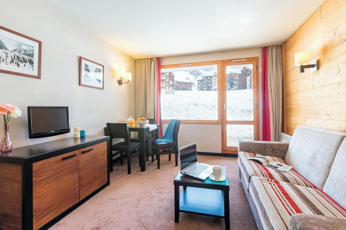 Une pièce chaleureuse avec un canapé, une petite table sur laquelle se trouvent un ordinateur portable et une tasse, un téléviseur posé sur un meuble, et une fenêtre donnant sur des bâtiments recouverts de neige.