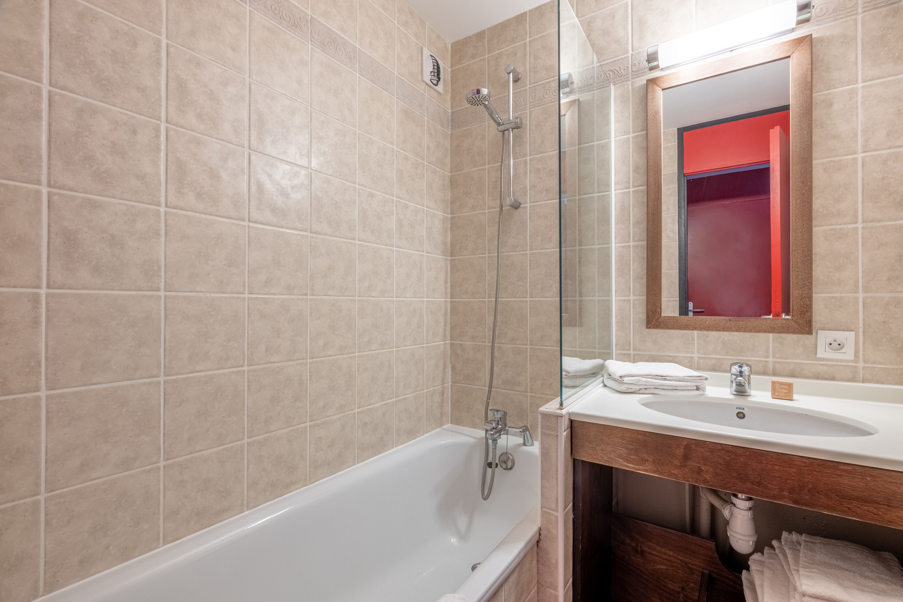 A modern bathroom with beige tiles, a white bathtub, a glass shower enclosure, and a wooden vanity with a sink and towel stack.
