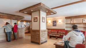 A reception area with wooden beams, a reception desk, and a seating area with a red sofa and a wooden coffee table. - Pierre & Vacances Residence Le Quartz (La Plagne-Tarentaise)