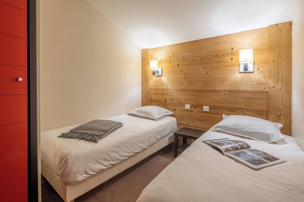 Une chambre avec deux lits simples, chacun équipé d’un oreiller et d’une couverture. Une petite table se trouve entre les lits. La chambre est dotée d’une tête de lit en bois et de deux lampes fixées au mur.