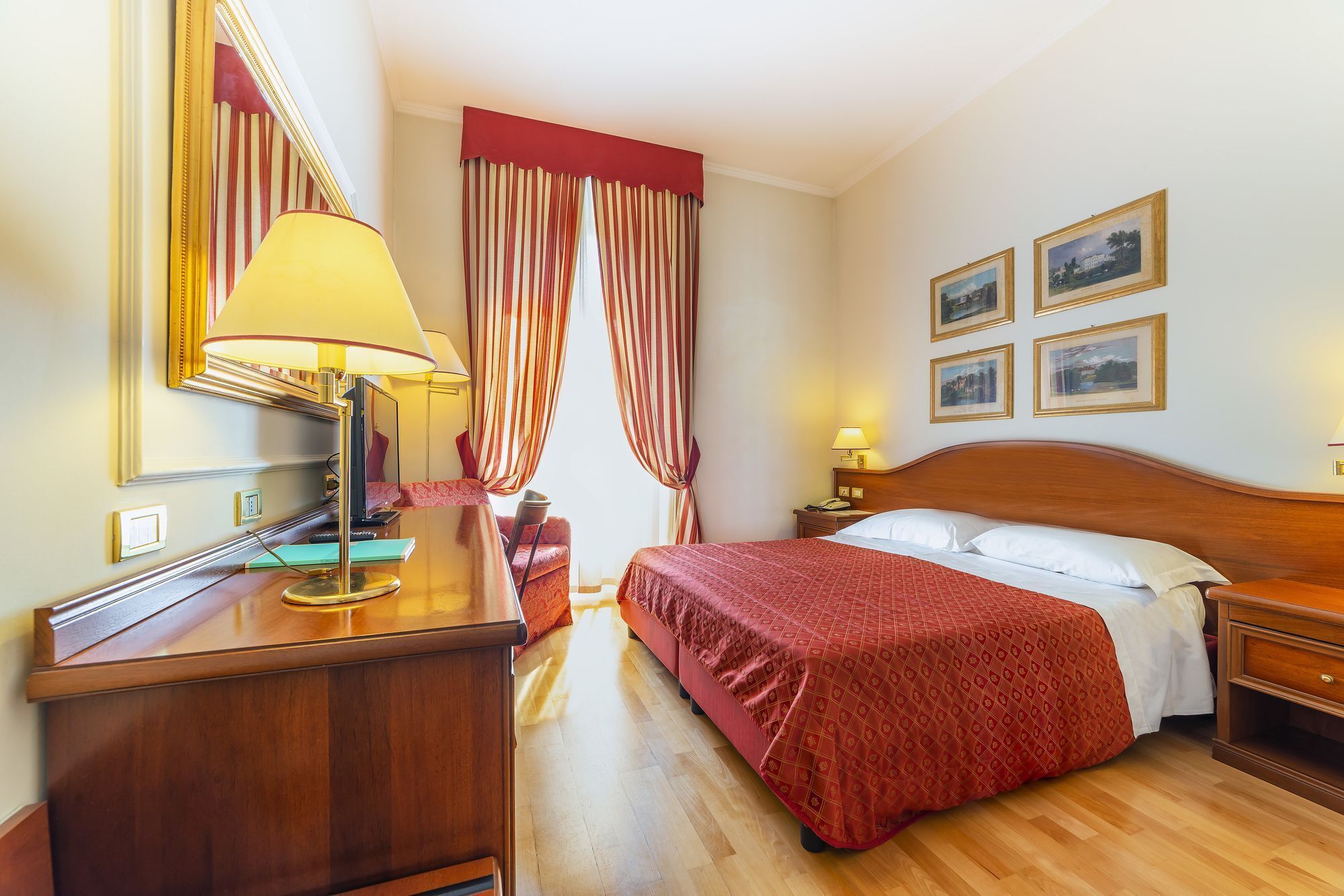A hotel room with a wooden bed, a desk with a lamp, a mirror, and a window with curtains.