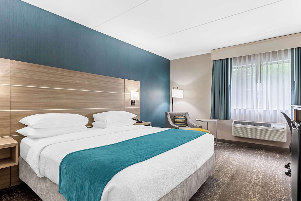 Una habitación de hotel con una cama grande, una mesita de noche, una silla, un escritorio, un ventanal con cortinas y un equipo de aire acondicionado.