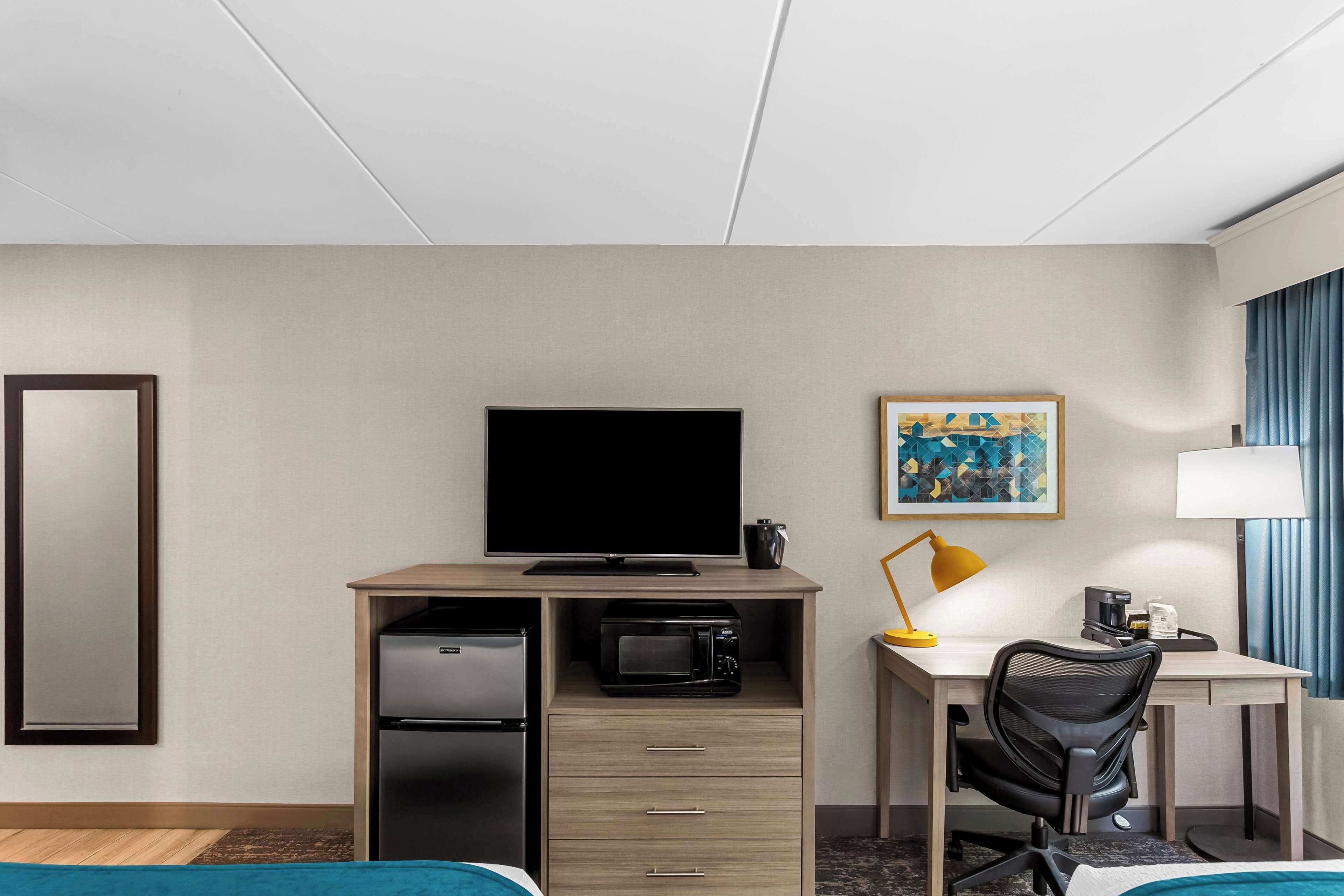 Habitación de hotel con cama, escritorio con computadora, televisor, microondas, cafetera y lámpara.
