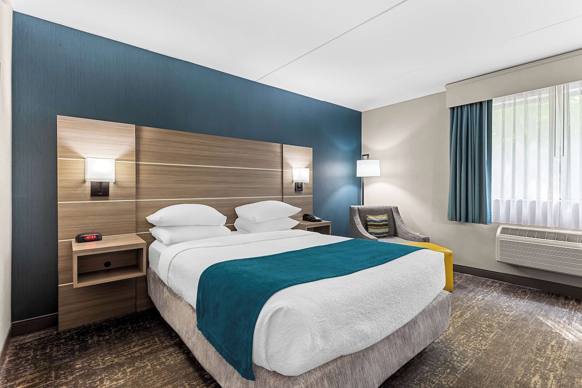 Una habitación de hotel con una cama grande, lámparas de noche, una mesita de noche, una silla y una ventana con cortinas.