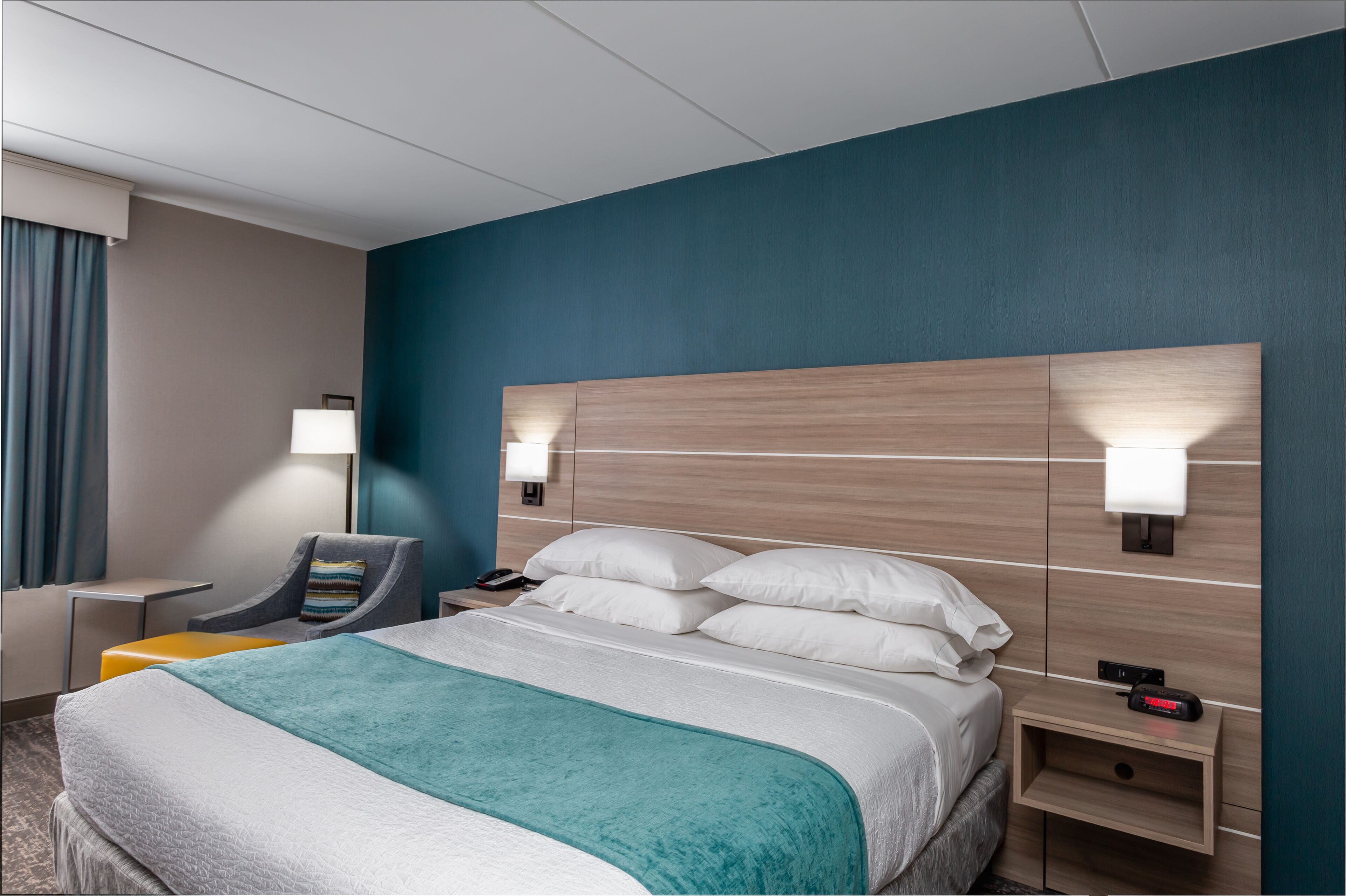 Una habitación de hotel con una cama grande, dos mesitas de noche, una silla y una mesita con una lámpara.