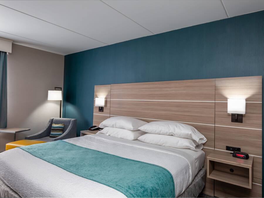 Una habitación de hotel con una cama grande, dos mesitas de noche, una silla y una mesita con una lámpara.