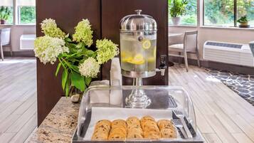 Mesa de buffet con dispensador de agua, bandeja de pasteles y un jarrón con flores.