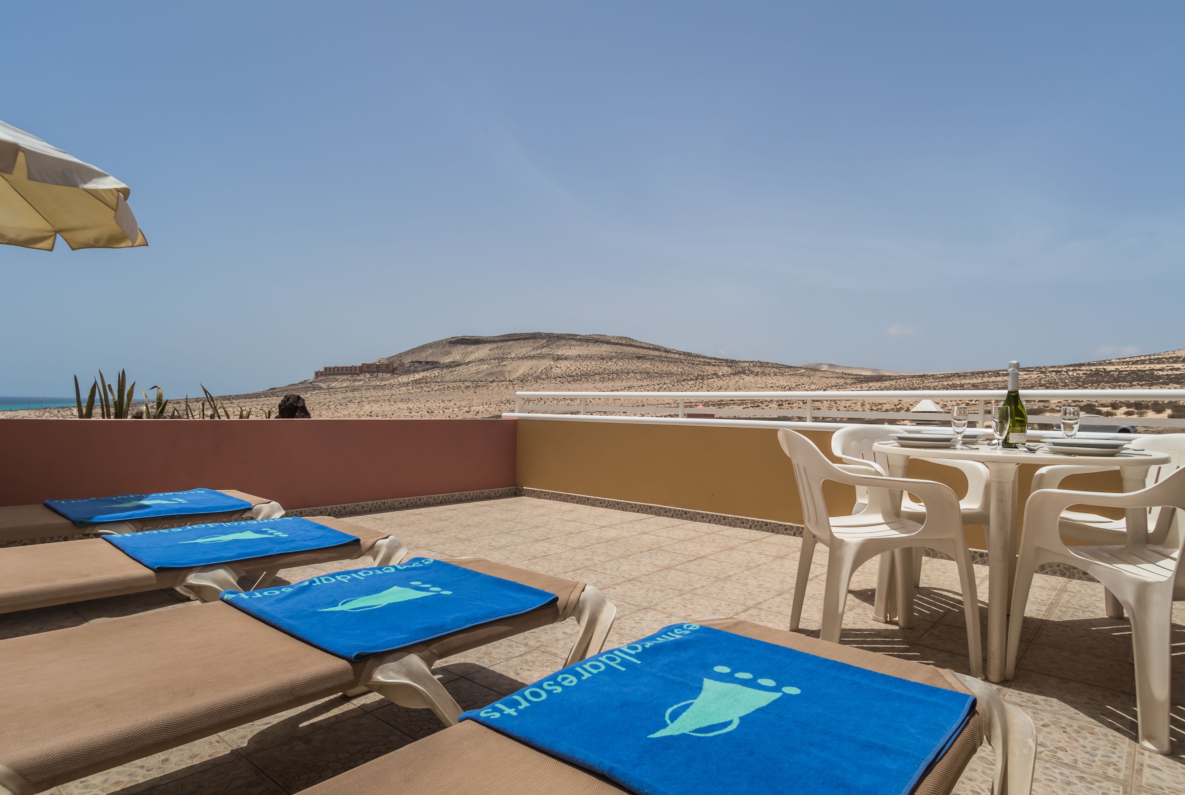 Eine Terrasse mit Liegestühlen und einem Esstisch, mit Blick auf einen Strand und ein Gebirge.
