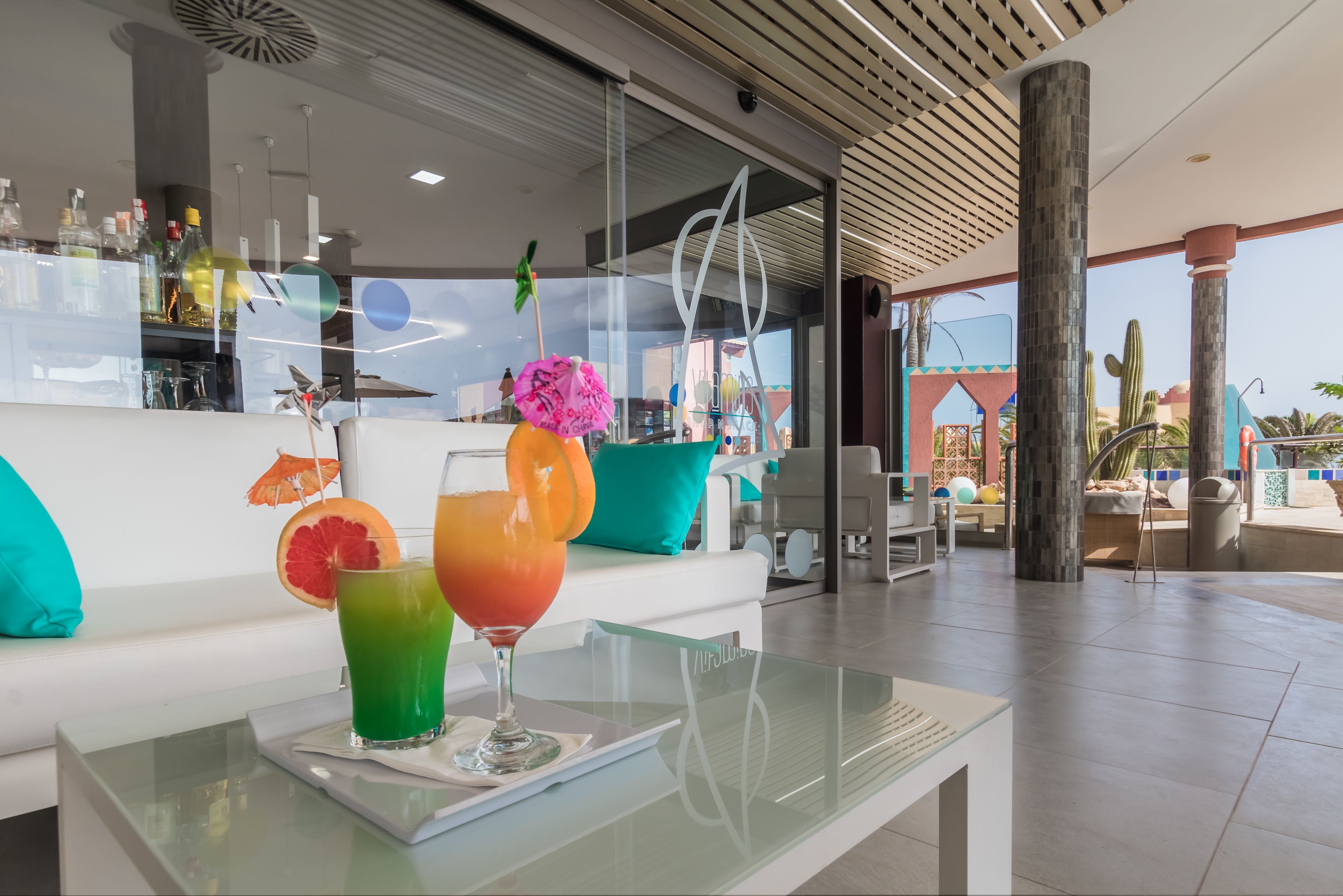 A modern bar area with a glass table displaying colorful drinks and a white sofa with teal cushions.