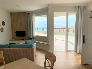 Een woonkamer met een televisie, een bank, een rieten stoel en uitzicht op de zee door een glazen deur.