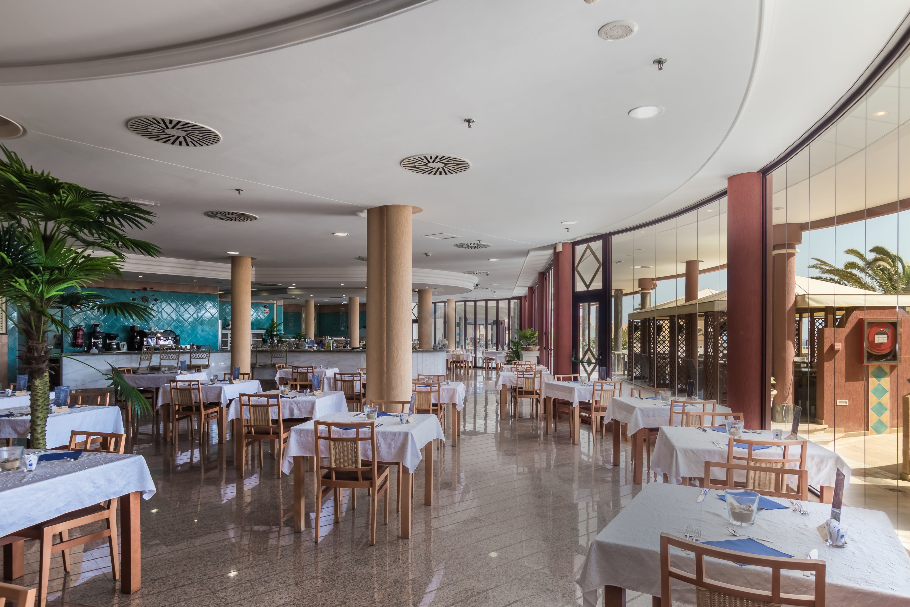 A spacious indoor dining area with multiple tables and chairs, large pillars, and a view of the outside through large windows.