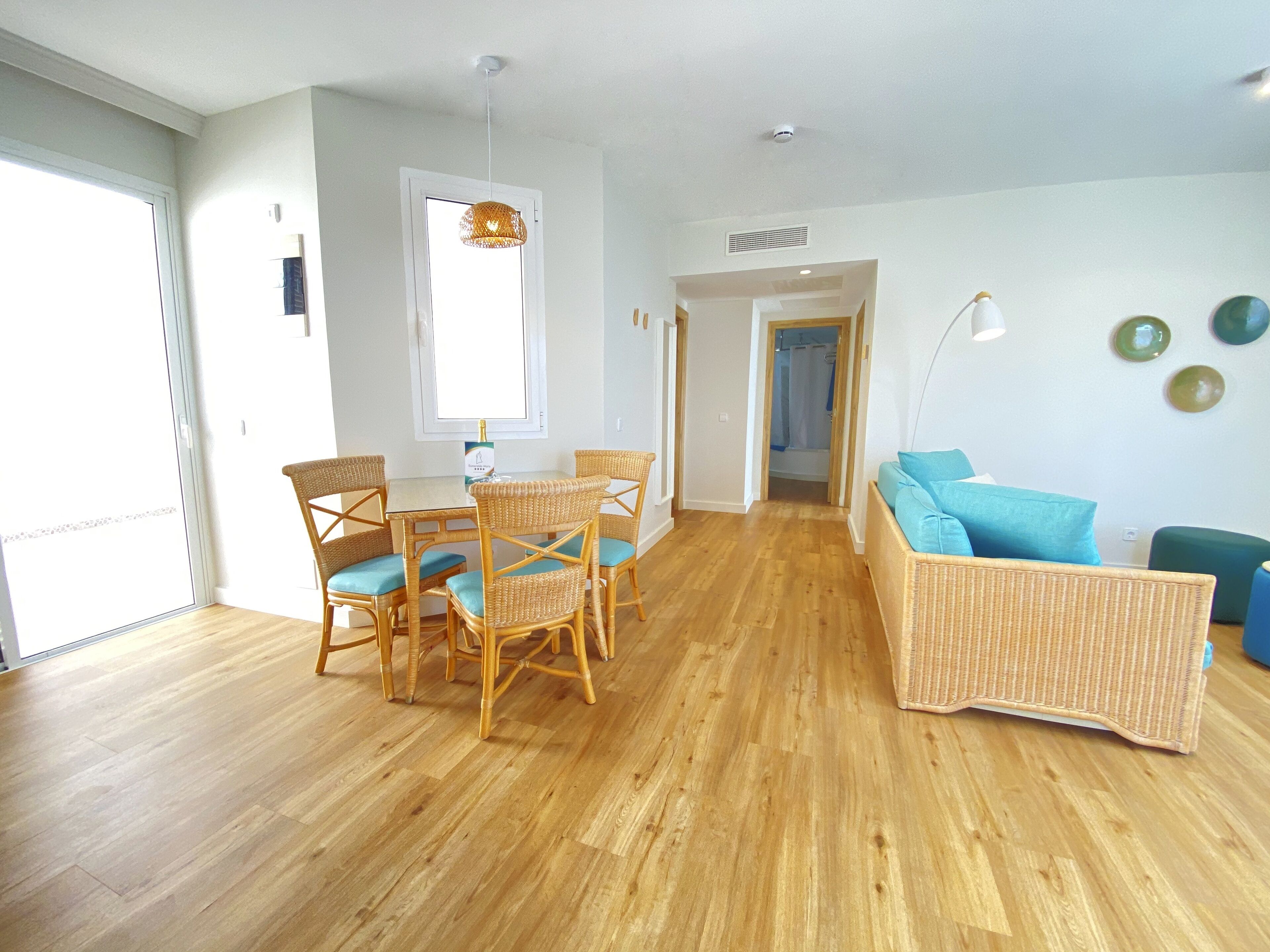A bright living room with a dining area, wooden flooring, wicker furniture, and a view through an open door.
