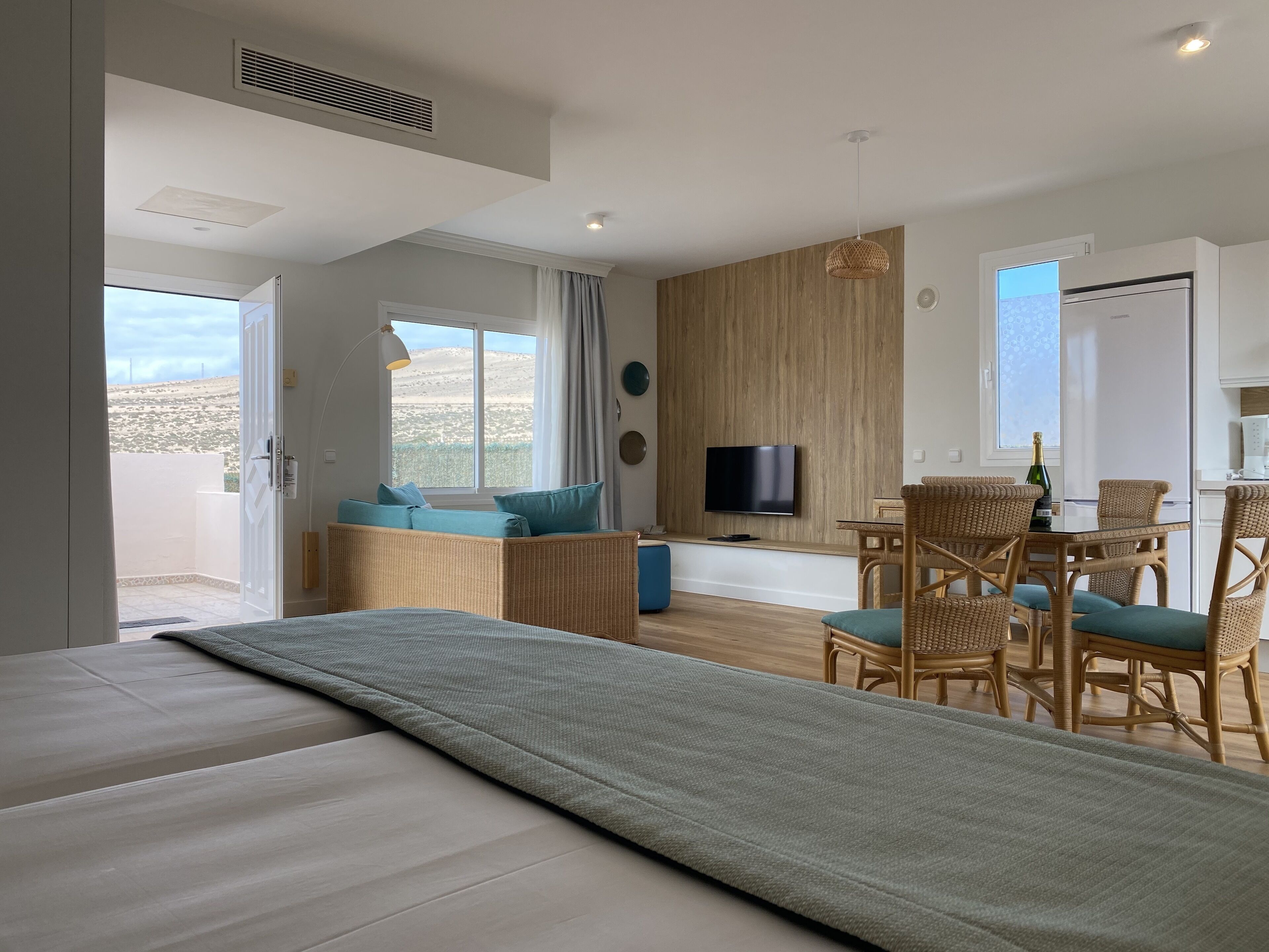 A modern hotel room with a bed, a seating area with cushions, a dining table with chairs, a TV mounted on the wall, and a view of the outdoors.