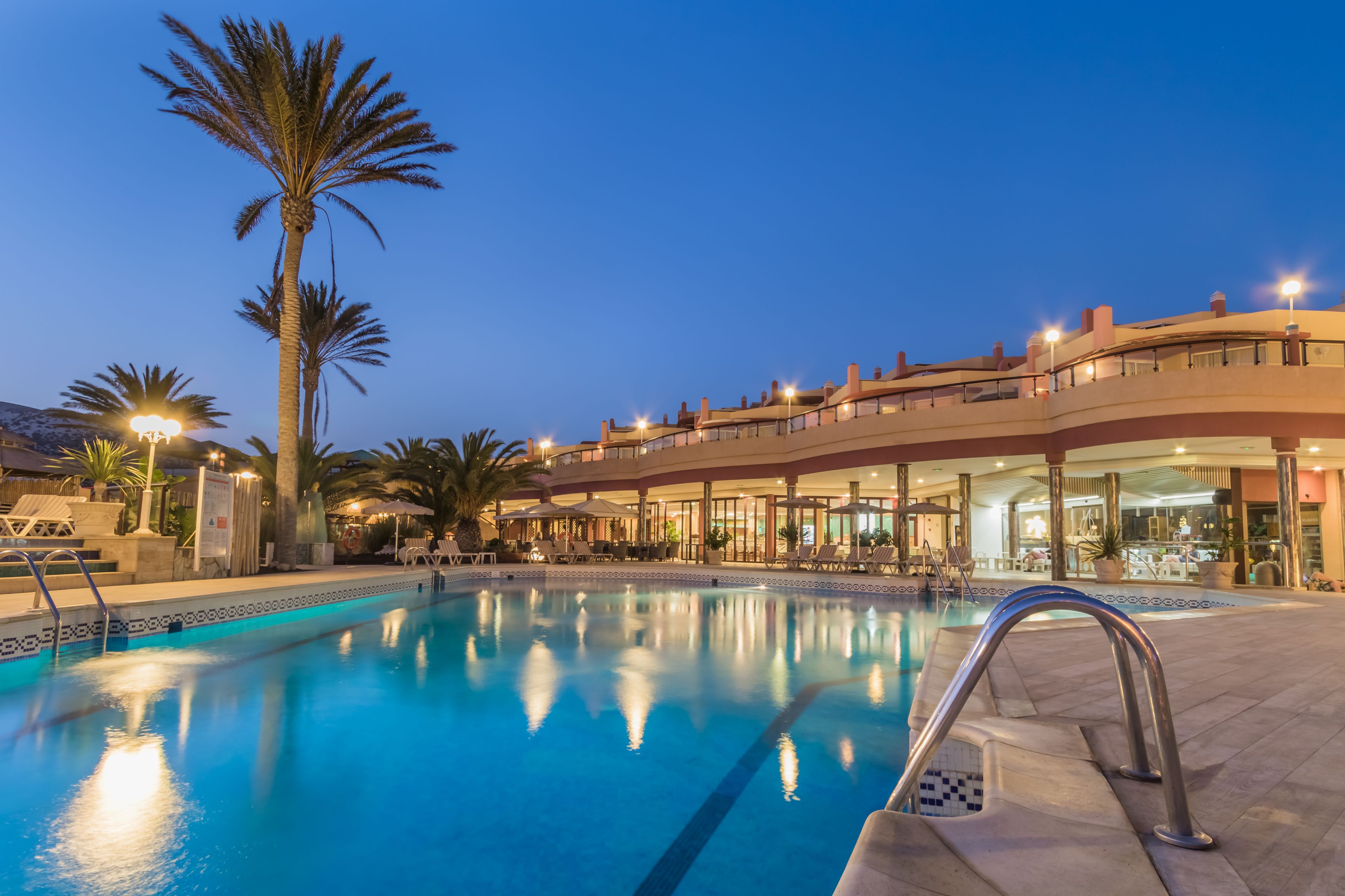 A swimming pool with a clear blue water surface, surrounded by palm trees and a building with multiple balconies and outdoor seating areas.