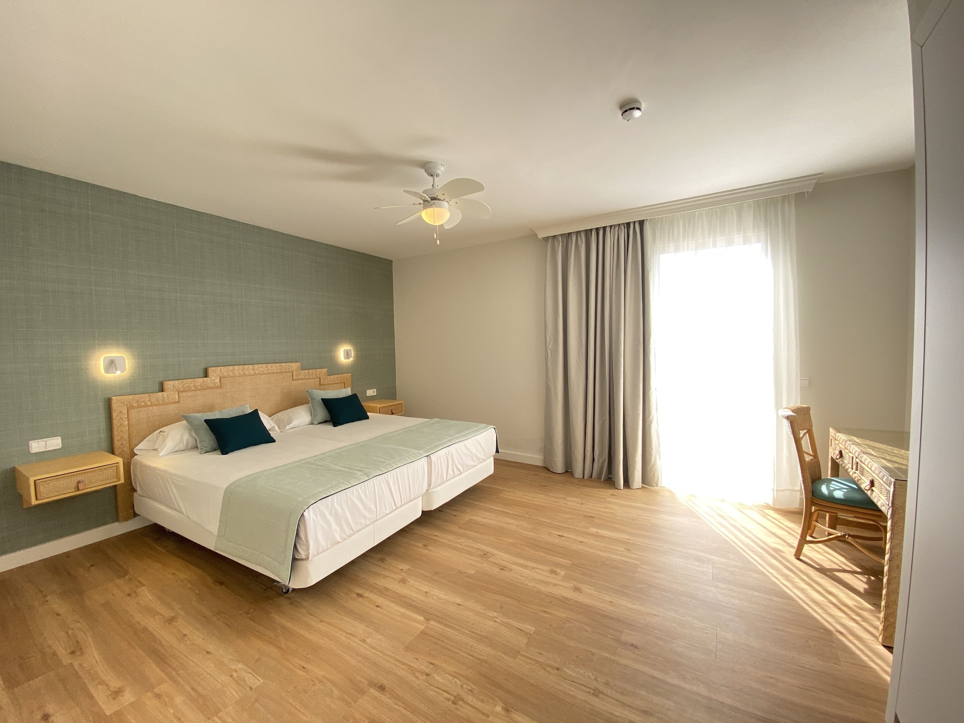 Camera da letto con un letto, pavimento in legno, ventilatore a soffitto e una finestra con tende.