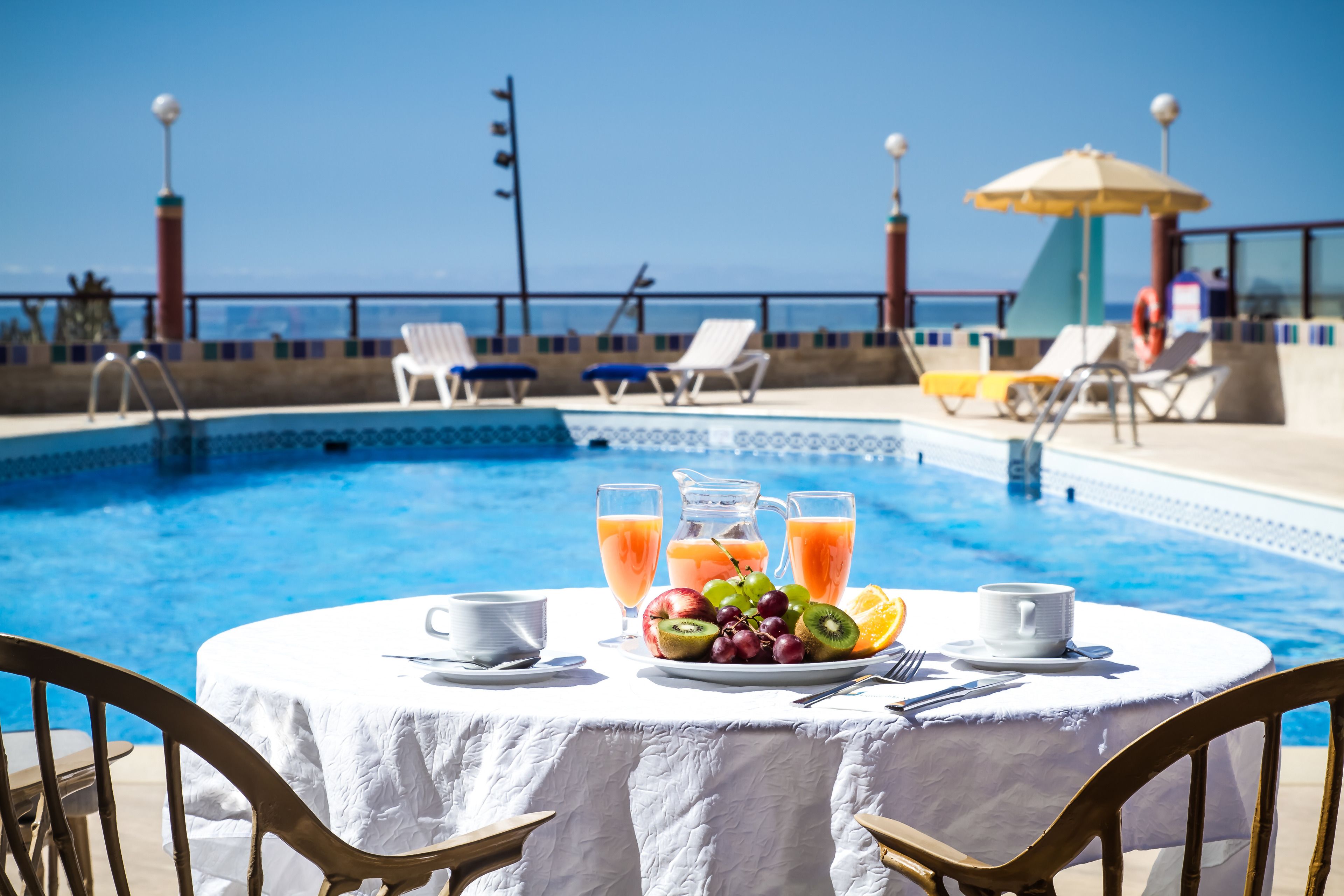A poolside dining area with a table set for two, a pitcher of juice, and a plate of fruit.