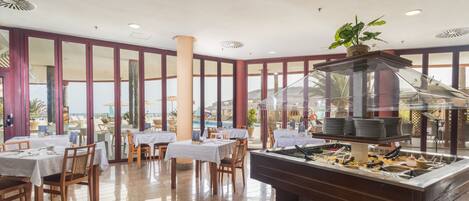 Un restaurante con mesas y sillas, una zona de bufé y grandes ventanales con vistas a la playa.