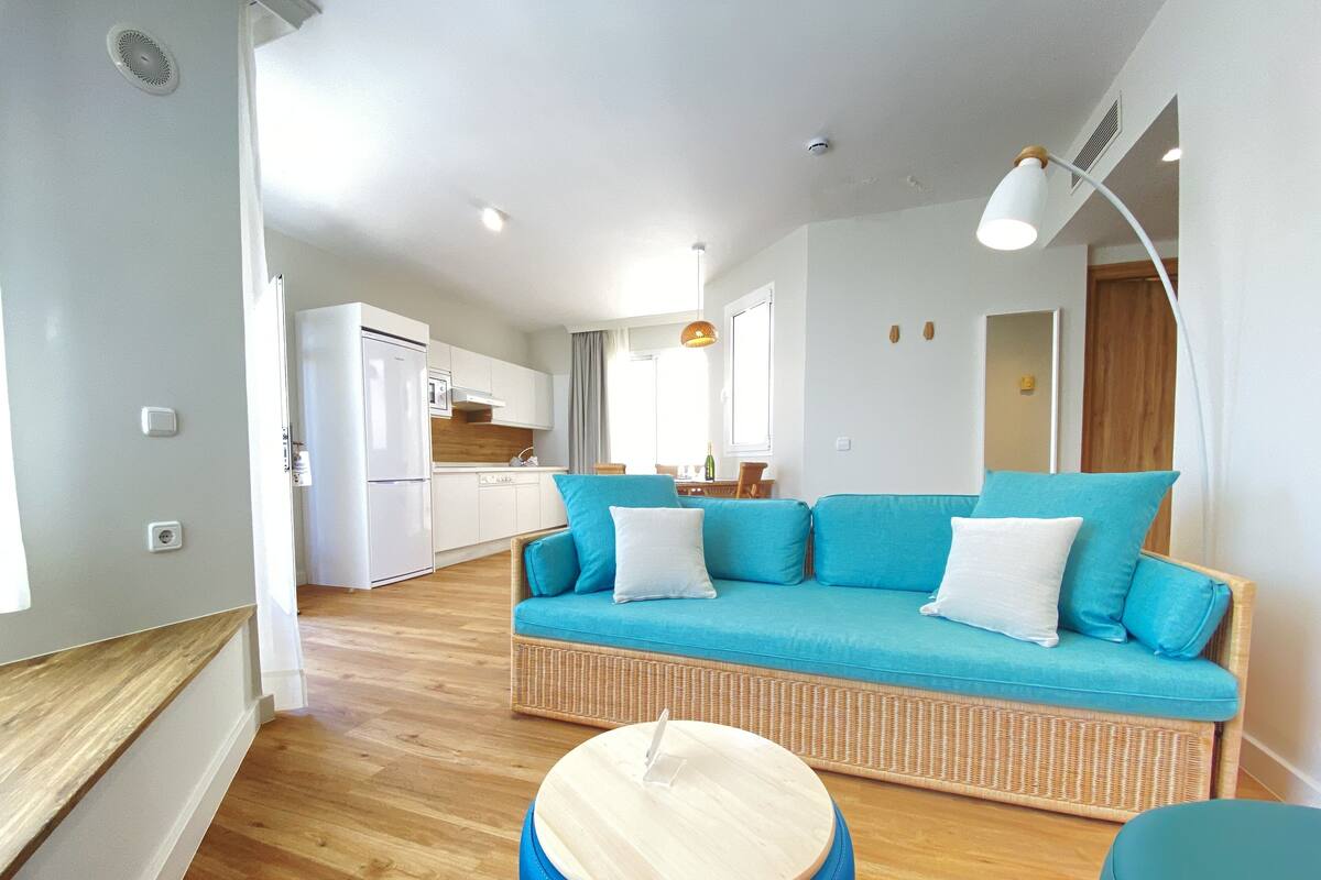 Un salon moderne avec un canapé bleu, un pouf en bois et une cuisine en arrière-plan.