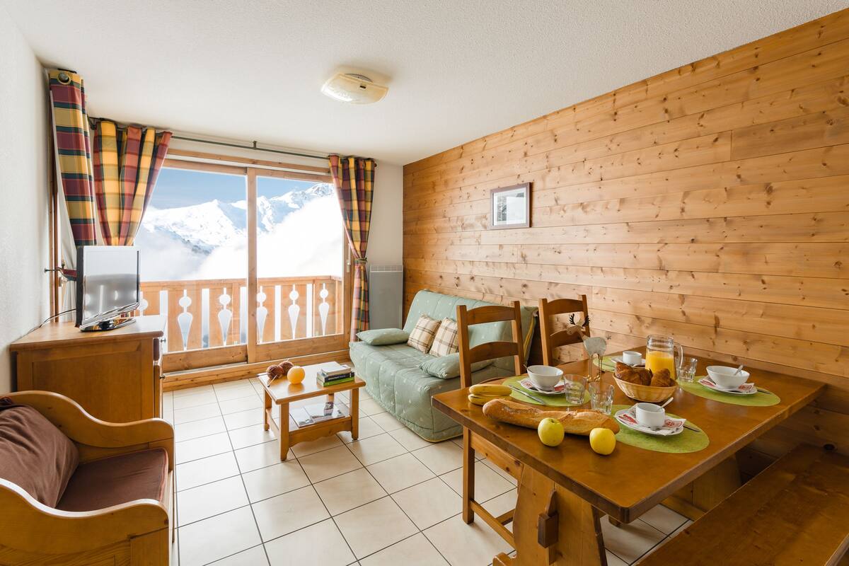 A cozy room with a wooden dining table set for breakfast, a sofa, and a TV on a wooden cabinet.