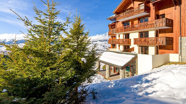 Eine Holzchalet mit Balkonen, einem Steineingang und einem verschneiten Berg im Hintergrund.