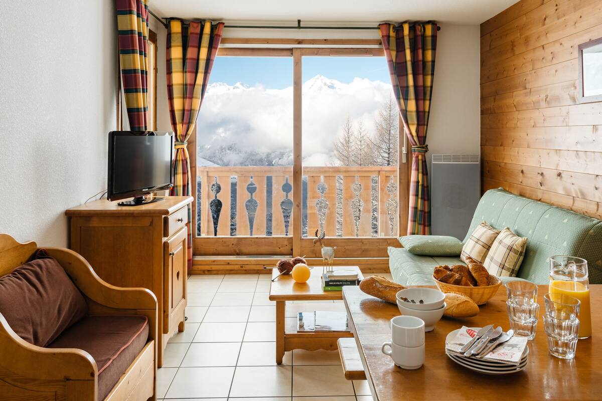 A cozy room with a wooden sofa, a dining table with breakfast items, a TV on a wooden cabinet, and a view of snow-capped mountains through a window with plaid curtains.