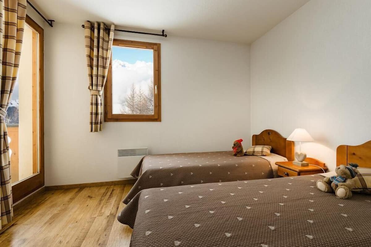 A room with two beds, a window with curtains, a nightstand with a lamp, and a teddy bear.
