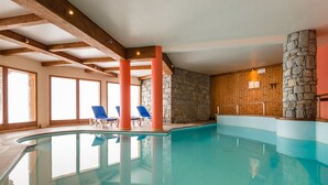 Ein überdachter Swimmingpool mit klarem Wasser, Holzbalken, Steinwänden und blauen Liegestühlen.