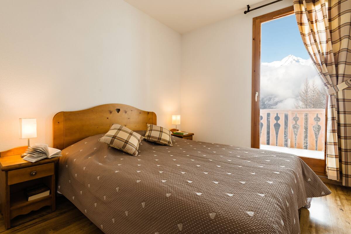 A bedroom with a wooden headboard, a bed with a patterned bedspread, two pillows, a nightstand with a lamp, and a window with curtains.