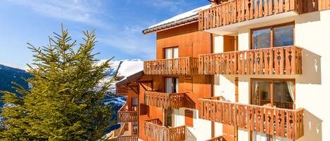 Un chalet in legno con balconi, un tetto innevato e un pino in primo piano.