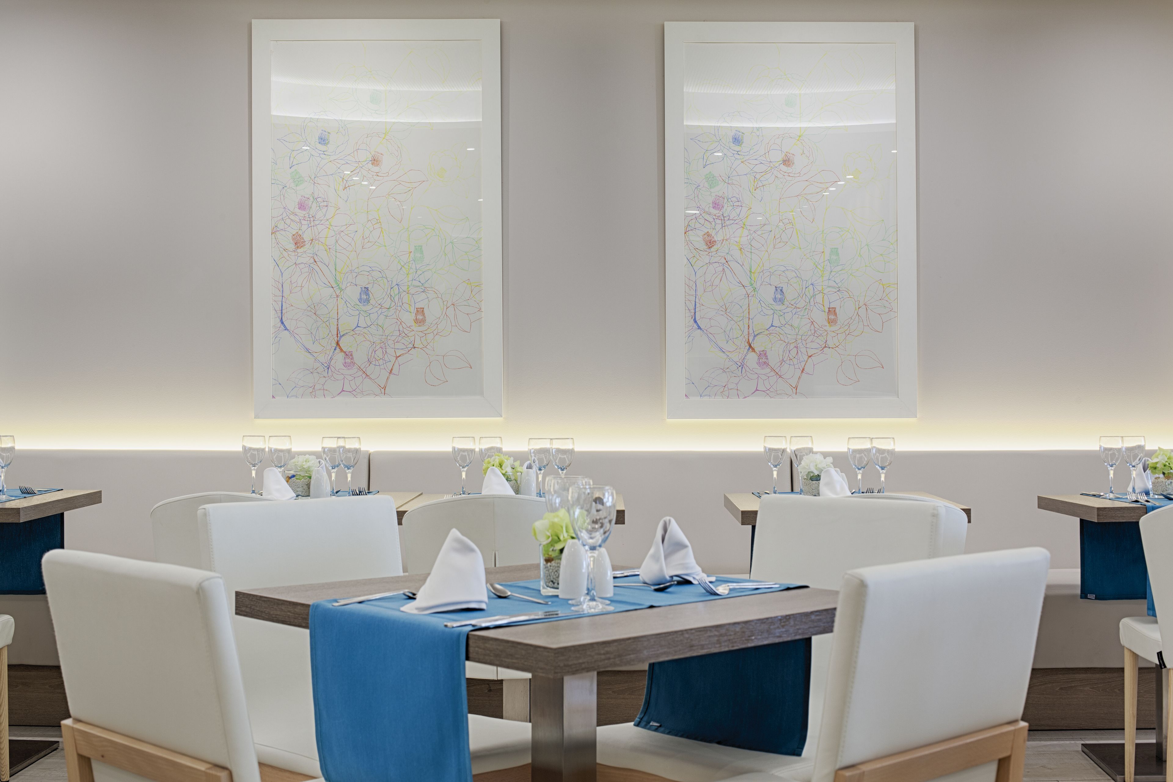 A dining area with tables set for four, each with a blue table runner, white chairs, and glassware.