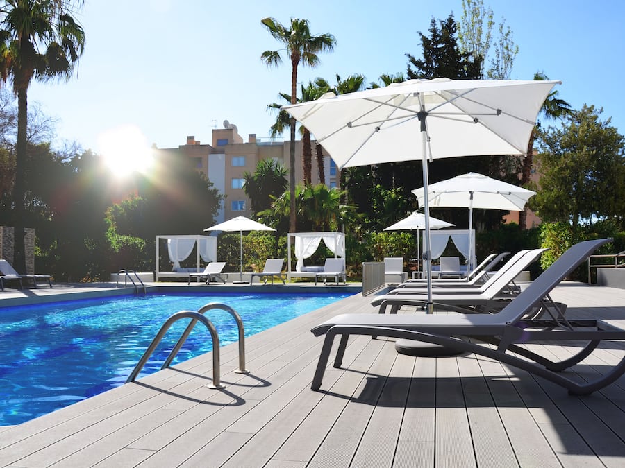 Un área de piscina con sillones de descanso y sombrillas, rodeada de palmeras y un edificio al fondo.