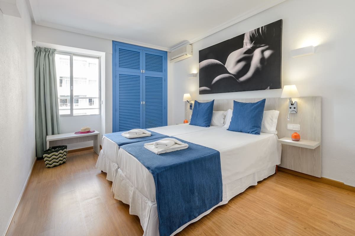 Habitación de hotel con una cama grande, cabecera azul, una obra de arte en la pared, mesita de noche con lámpara y una ventana con cortinas.
