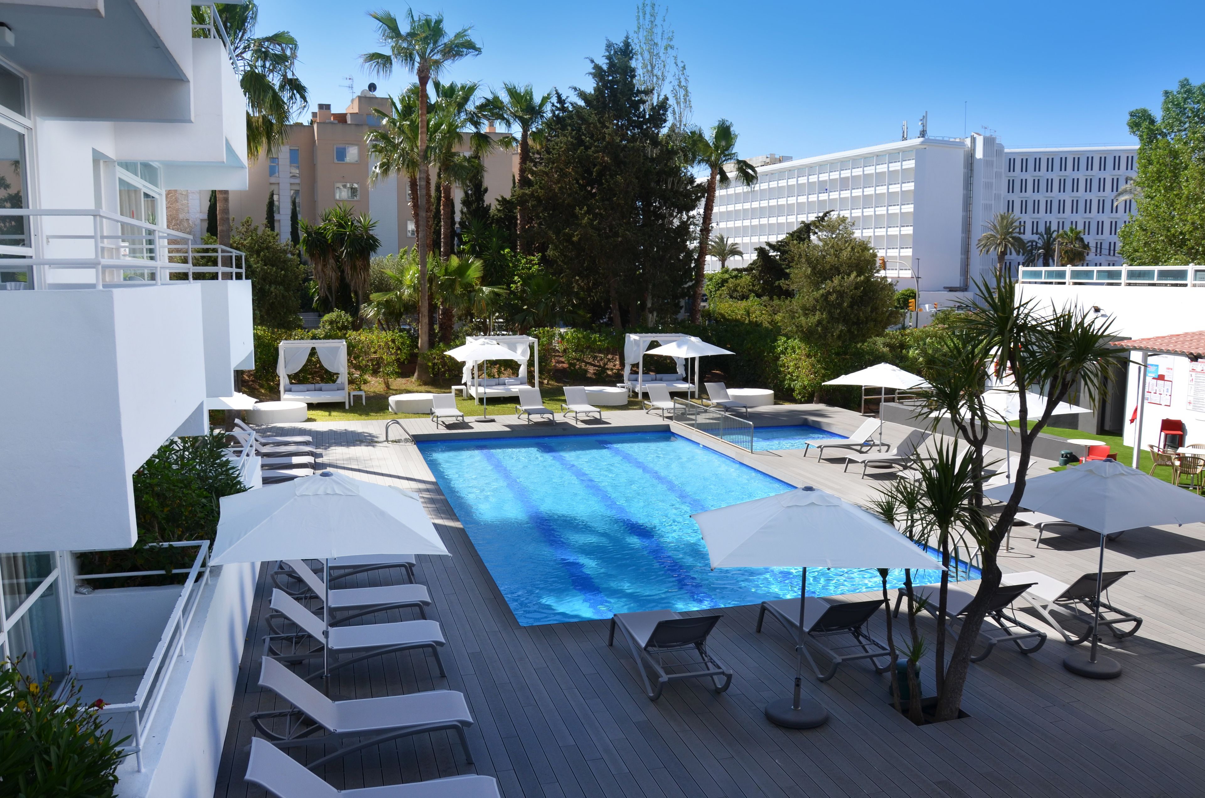 Une piscine entourée de palmiers et d’immeubles, avec des chaises longues et des parasols.
