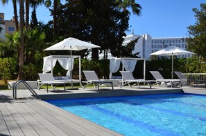 Ein Pool mit weißen Liegestühlen und Sonnenschirmen, umgeben von Palmen und einem Gebäude im Hintergrund.