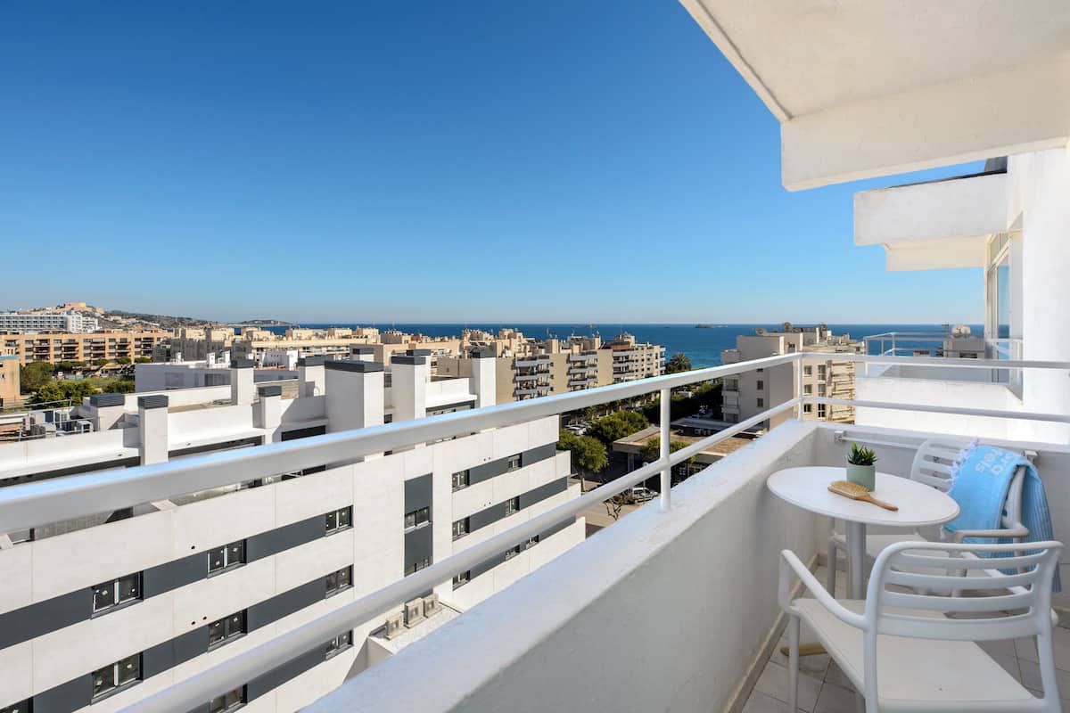 Un balcón con vistas a la ciudad y al mar, muebles blancos y una pequeña mesa con una planta.