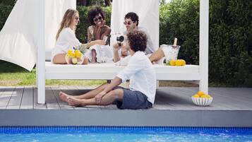 Escena junto a la piscina con una pérgola blanca, una piscina y una mesa con frutas y bebidas.