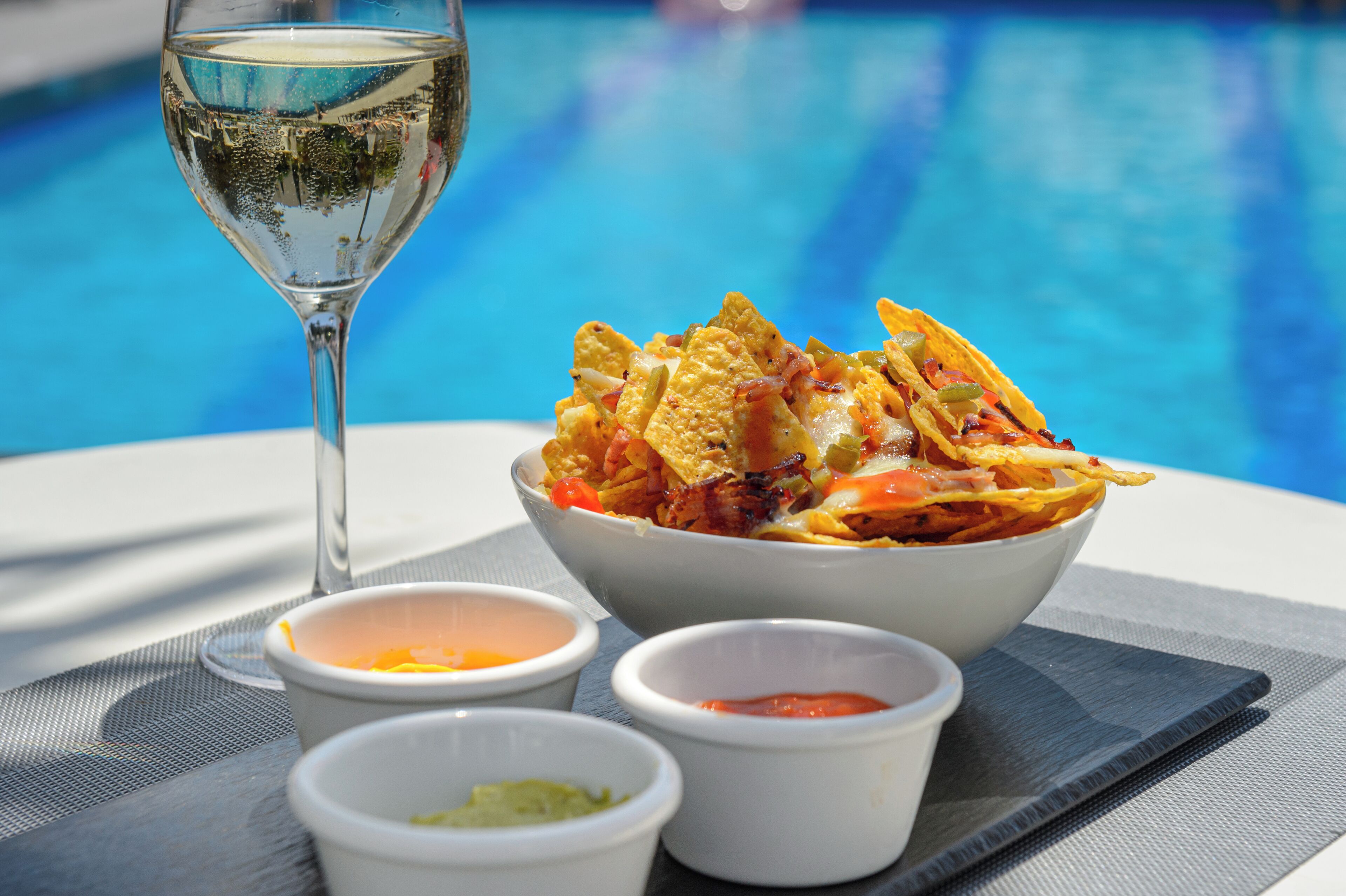 Un plato de nachos con tres salsas para mojar y una copa de vino blanco junto a una piscina.