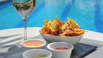 Un plato de nachos con tres salsas para mojar y una copa de vino blanco junto a una piscina.