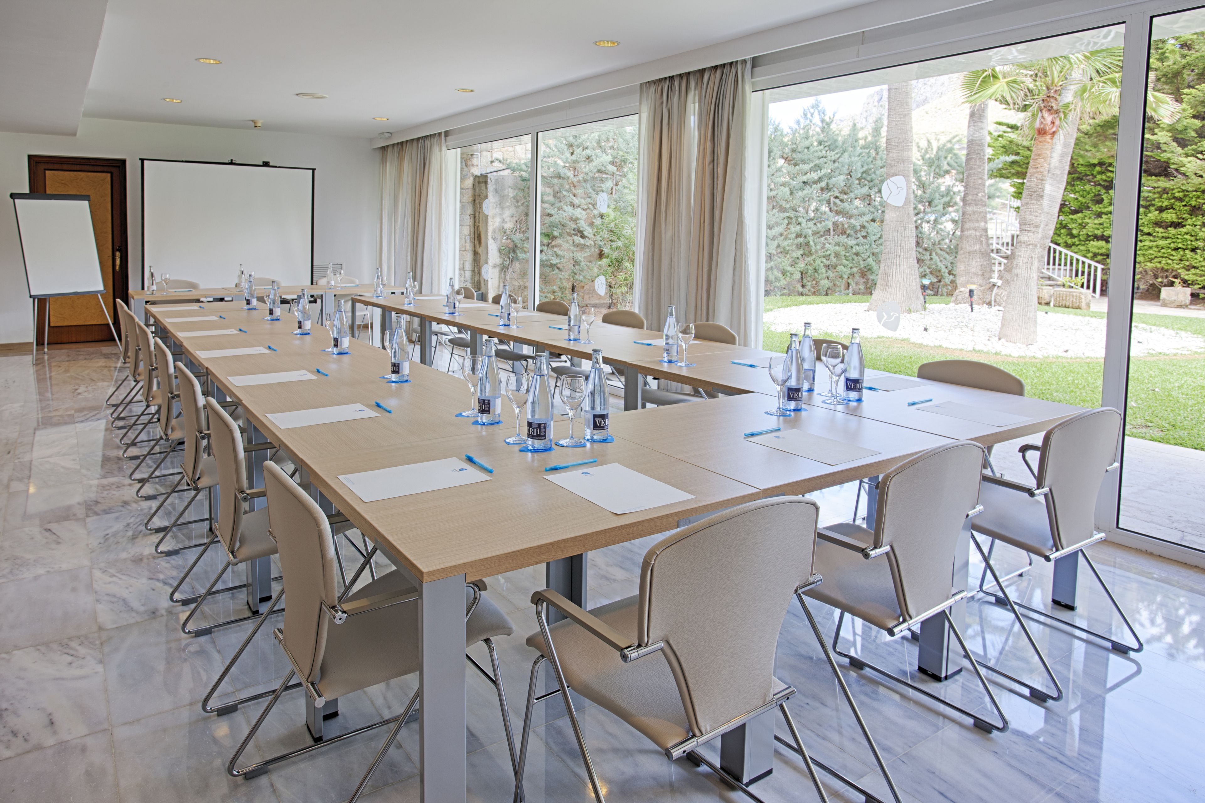 Un salón de conferencias con una larga mesa, sillas, una pizarra y ventanas amplias con vistas a árboles y un sendero.
